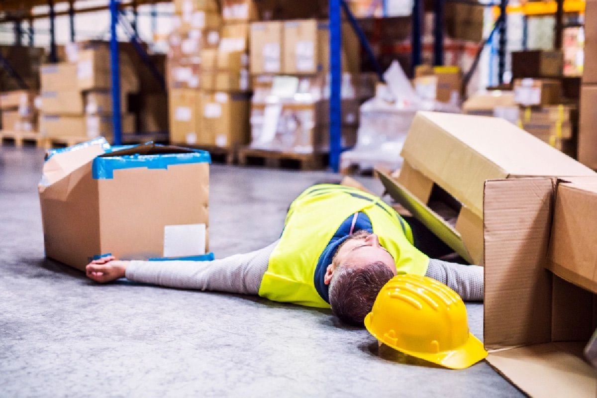 Man in high-visibility vest unconscious on warehouse floor with hard hat nearby.